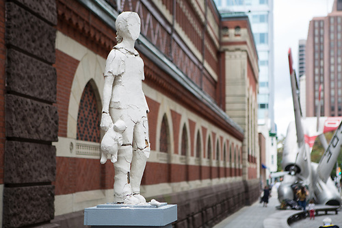 Photograph of clay sculpture of a young girl on pedestal with streetscape in background.
