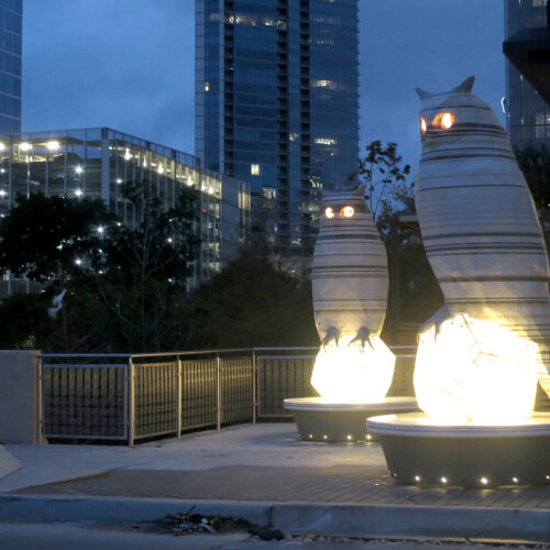 Photograph of owls sitting on pillars lit from below at night time.