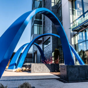 A blue sculpture with three arches, positioned in front of a building