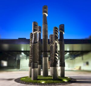 A group of illuminated tall, cylindrical sculptures with textured surfaces against a blue evening sky