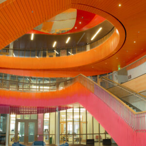 This interior image showcases a modern, vibrant architectural space featuring sweeping orange and pink staircases with curved design elements. The staircases are complemented by sleek railing designs and are illuminated by recessed lighting, creating a warm and inviting atmosphere. The space includes a lounge area with comfortable blue armchairs arranged around small circular tables, positioned on a bright red carpet. Large glass windows and doors provide a view into the adjacent rooms or offices, enhancing the openness and accessibility of the area. The design emphasizes dynamic geometry and bold color choices, making the environment lively and contemporary.