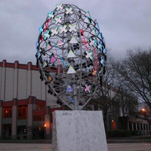 This image depicts a tall, modern sculpture resembling an egg shape, composed of a metallic framework decorated with various colorful, illuminated geometric shapes, including triangles, crosses, and other abstract symbols. The sculpture is situated outdoors on a white pedestal, with a late afternoon or early evening sky overhead. Behind it, there is a large building with a distinctive red and grey industrial-style façade, and on the right side of the image, there are leafless trees, indicating it might be late fall or winter. The overall scene has a contemporary, artistic atmosphere with a somewhat overcast sky.
