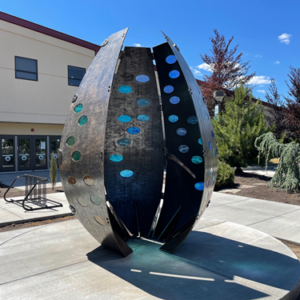 Metal ovular sculpture is opening is position on a circular concrete plaza. Ovular cutouts are filled with teal and blue glass inserts. A building sits in the background.