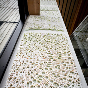 This image shows four intricately designed lace or crochet-style pieces laid out on a long white table. The designs are delicate and detailed, resembling natural patterns such as coral or organic cellular structures. The table is positioned next to a window with a wooden frame, and the setting appears to be a modern, well-lit interior space with wooden paneling on the ceiling and part of a glass display case visible on the right. The focus is on the textured, artistic fabric pieces arranged neatly in a row.