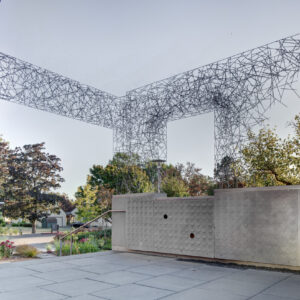 The image shows a modern outdoor installation with a distinctive metal framework structure that appears to be a sculpture or architectural feature. The structure is composed of thin, intersecting metal rods forming a geometric, lattice-like pattern. It extends horizontally and then rises vertically in an angular fashion, creating a visually striking form that somewhat resembles abstract wings or a stylized shape. In the foreground, there's a wall with a textured, circular pattern design, and the area around the installation is landscaped with trees, plants, and flowers. The setting appears to be an urban or cultural space, possibly a sculpture park, plaza, or art center. The sky is clear, suggesting it is daytime, and the overall atmosphere is calm and clean.