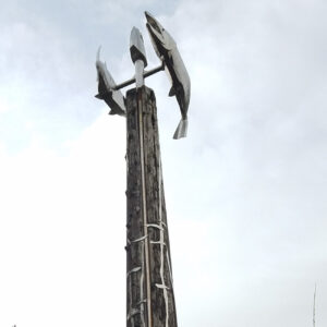 A 40-foot vertical pole created from a salvaged log with a steel and cast framework that depicts the salmon perched atop. Surrounded by wooded area.