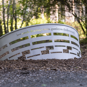 This image shows a metallic, curved memorial or sculpture placed on the ground in a wooded area. The structure features horizontal cutouts and has an inscription written in a foreign language, which appears to be Turkish. The background consists of trees with sunlight filtering through their leaves, creating a peaceful and contemplative atmosphere. The ground around the sculpture is bare soil with some small plants, adding to the natural setting.