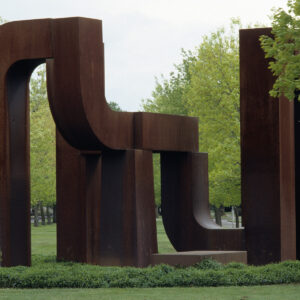 This image features a large, abstract sculpture located outdoors in a park-like setting. The sculpture is composed of several massive, rust-colored metal pieces with bold, geometric shapes and flowing curves. It appears to be a modern or minimalist art piece, with elements that resemble both organic and industrial forms. Surrounding the sculpture, there is lush green grass and trees, contributing to a peaceful and natural environment. The background shows a clear sky and a quiet street, indicating that the sculpture is situated in an open, serene area.