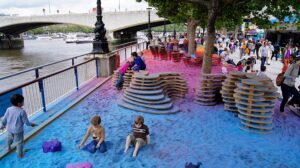 Children playing in colorful sand among wooden structures on a riverside walkway.