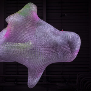 This image features a large, abstract, three-dimensional art installation suspended from the ceiling in a dimly lit indoor space. The sculpture has a soft, organic shape with rounded bumps and a smooth surface, illuminated with warm hues of yellow, orange, and pink. The lighting creates a glow that enhances the textures and contours of the piece. Additionally, the installation casts a vibrant, intricate reflection or shadow onto the wall behind it, displaying a pattern of small, bright dots and lines that mirror the sculpture's form, adding to the visual complexity of the scene.