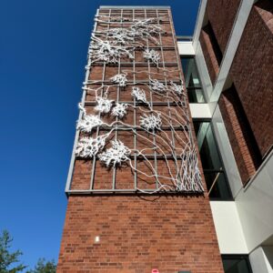 This image shows the exterior of a modern brick building with a tall, narrow vertical window in the center. On the brick facade, there is an artistic installation resembling white, branching tree roots or lightning extending across the front of the building. The sky is clear and blue, and there are some green trees and plants along the sidewalk at the bottom of the image.