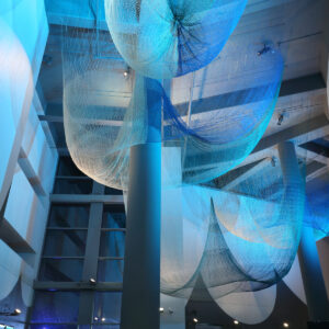 This image depicts an interior space with an artistic or modern architectural design. The ceiling features large, flowing, mesh-like fabric structures in shades of blue, creating a dynamic and textured visual effect. The space is illuminated with cool lighting, enhancing the serenity and contemporary aesthetic. The structure is supported by multiple pillars, and there are visible large windows or glass panels that suggest a spacious and open atmosphere. The overall ambiance is sleek, artistic, and sophisticated.