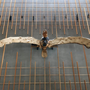 The image shows a mounted wooden sculpture of a large bird, likely an eagle, with its wings spread wide. The sculpture appears to be attached to a wall with a modern, geometric wooden framework. The bird has a detailed form, with a prominent beak, and the sculpture's materials seem to include carved and possibly painted wood. The background wall is plain and dark, contrasting with the lighter color of the sculpture. The setting looks like a modern interior space, possibly a museum or an art installation.