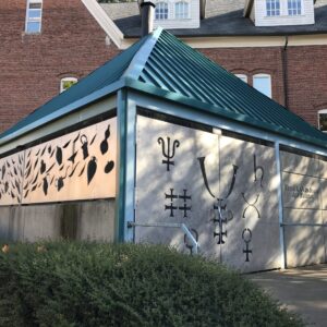 This image shows a small building with a green metal, pitched roof. The building has decorative metal artwork on its exterior walls, featuring various symbols, some of which resemble alchemical or astrological symbols, as well as abstract shapes and patterns. The background includes a larger brick structure with multiple windows, and there are bushes in the foreground. The overall scene appears to be in an urban or suburban setting.