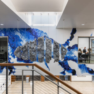 This image shows an indoor space with a modern, striking mural on the wall. The mural features an abstract design with a prominent central element resembling a textured, honeycomb-like structure in shades of gray or black. Surrounding this are vivid blue splashes and streaks that create an energetic, fluid backdrop. To the right, there is a glass-enclosed room where several people are seated, possibly in a waiting or meeting area. The space has a clean and contemporary aesthetic, with a staircase railing visible in the foreground and natural light illuminating the scene.