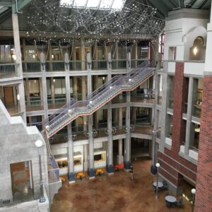 This image shows the interior of a modern, multi-story building. The area features a spacious atrium with a high ceiling and large skylights that allow natural light to illuminate the space. There are multiple levels with open walkways and railings, visible on the upper floors. A prominent staircase with a zigzag design connects the different floors, positioned centrally in the image. The decor includes brick walls and large windows. On the ground floor, there are a few small tables and chairs, and some decorative lighting fixtures, including streetlamp-style lamps. The overall aesthetic combines industrial and modern elements.