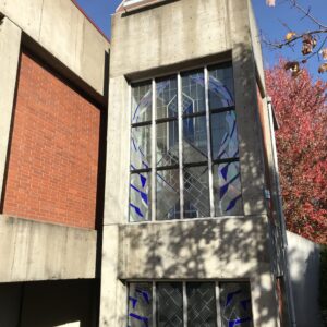 This image shows a modern, multi-story building with a striking architectural design. The facade features concrete walls, large stained glass windows with blue and purple accents, and a section resembling a clock tower at the top. The clock face is minimalistic, with only hour markers and a black minute hand, but no visible hour hand. The building is situated next to trees, with one tree casting shadows on its surface. The overall scene suggests it is a clear, sunny day.