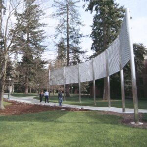 This image shows a park or outdoor area with a group of four people walking along a curved pathway. On the right side of the pathway, there is a large, modern, curved metal structure that is the art installation. The surrounding area is lush with green grass, and there are tall trees with some branches without leaves, indicating it might be early spring or late fall. In the background, there is a building with multiple windows, adding to the impression of an institutional or campus setting. The weather appears to be clear with sunshine, casting shadows on the ground.