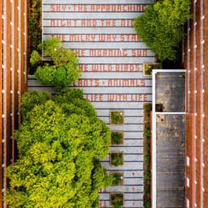 The image depicts an aerial view of a rectangular courtyard enclosed by tall brick buildings with multiple windows. In the center, there is a paved area with a pattern of rectangular tiles and some small planters with greenery. Large leafy trees are prominently situated within the courtyard, adding lush green contrast to the stone and brick surroundings. On one of the tiled sections, there is a message written in bold red letters that reads: "THE OCEAN + THE BLUE SKY + THE APPROACHING NIGHT AND THE DICK THE MICKEY DAY STAR THE MORNING SUN THE WILD BIRDS + ANIMALS LIFE WITH LIFE" The overall scene blends urban architecture with natural elements, creating a peaceful and inviting outdoor space.