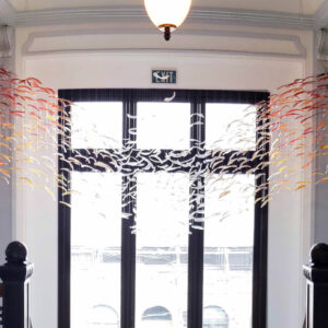 This image features a decorative installation inside a well-lit room with a large window and a black door at the center. The installation depicts a large tree with numerous birds flying around it, crafted with what appears to be metal or paper and hanging from the ceiling with strings, creating a dynamic scene. The railing in front suggests this is on an upper level or balcony. The overall ambiance is artistic and elegant, with the birds and tree adding a nature-inspired touch to the interior space.